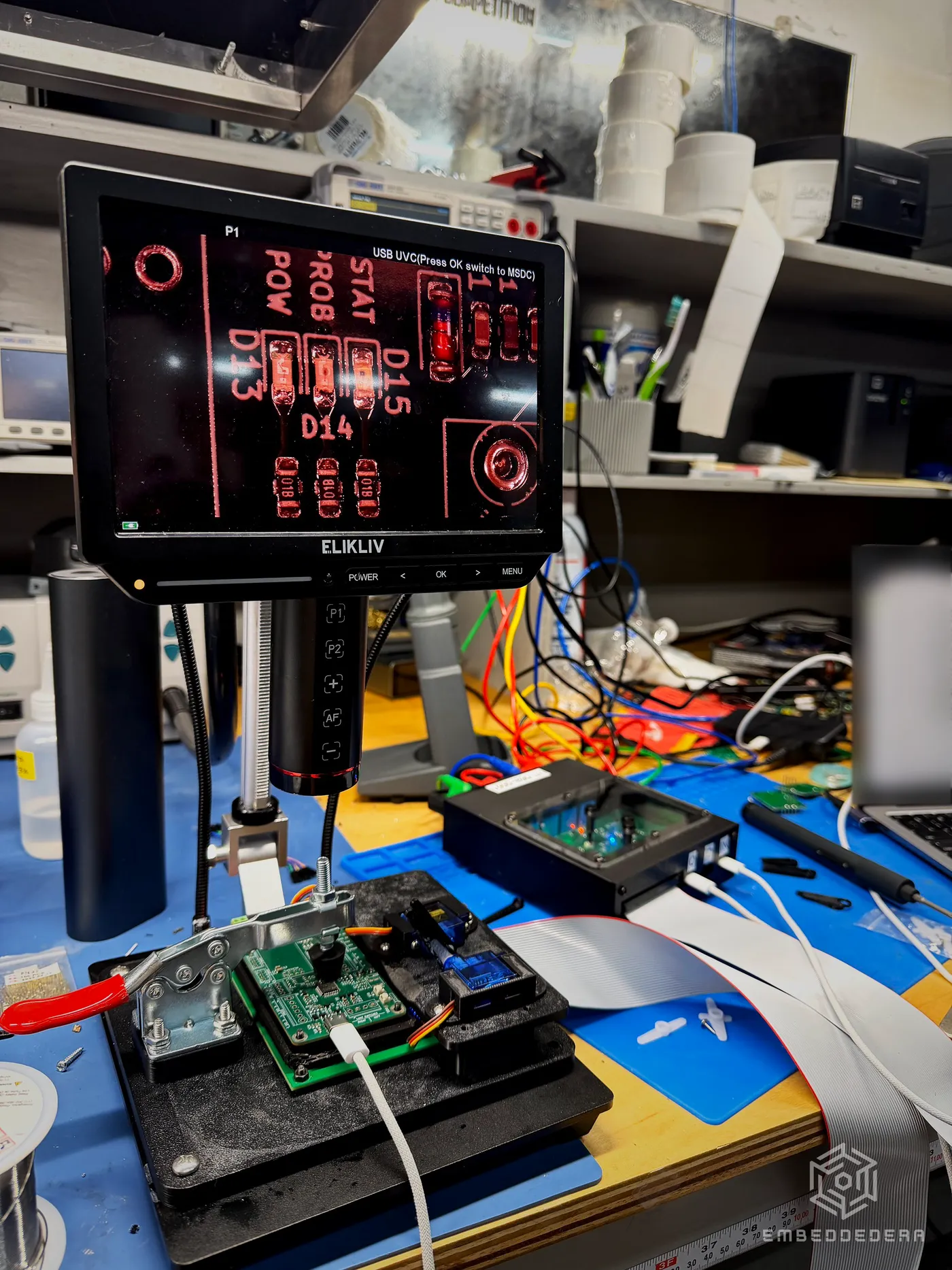 Close view of the camera head mounted over a test fixture, with a board clamped below it and a monitor above showing the camera’s view.