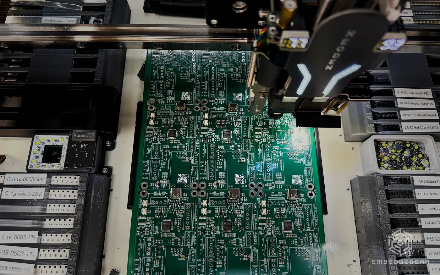 The pick-and-place head over a panel of EmbeddedEra boards, component feeders labelled and racked around it.