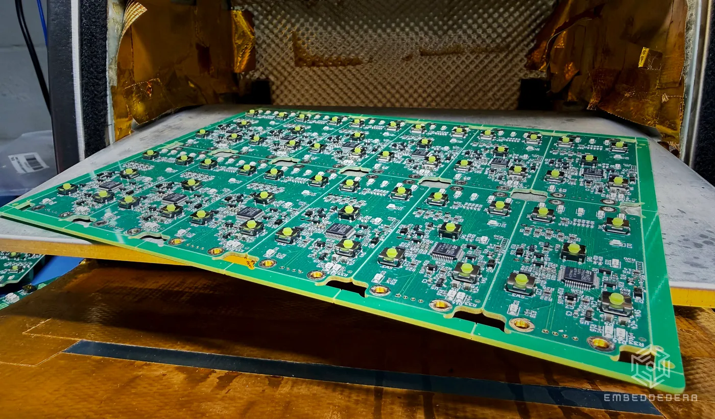 A freshly soldered panel of EmbeddedEra boards lifted in our open reflow oven, gold heat shielding behind it.