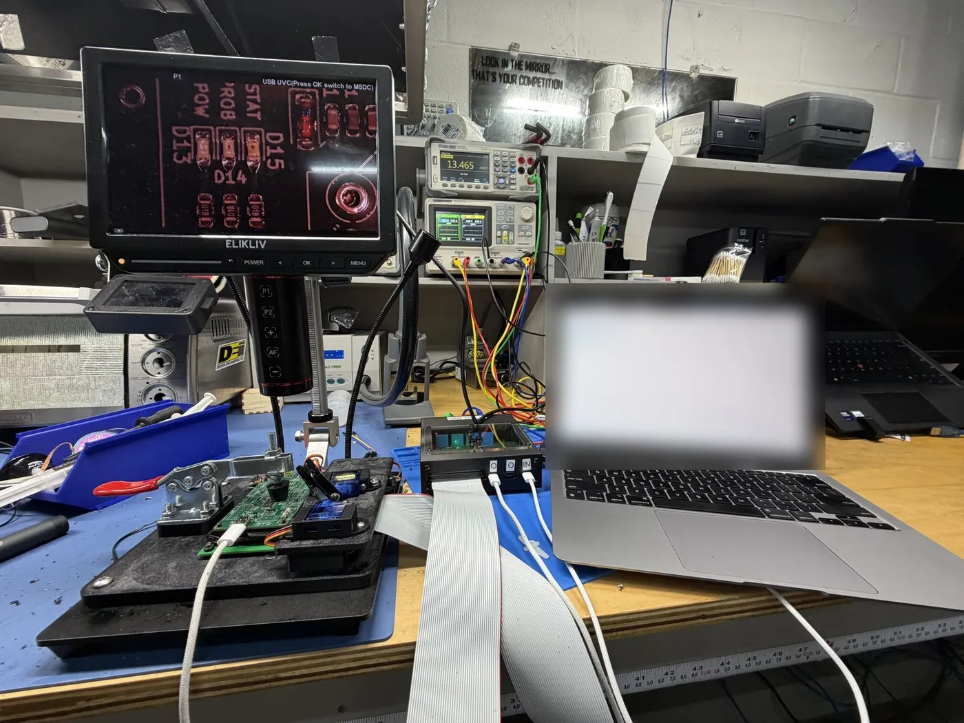 Our test station on the bench: a camera on a stand over the board under test, a monitor showing its view, bench supplies and a laptop behind.
