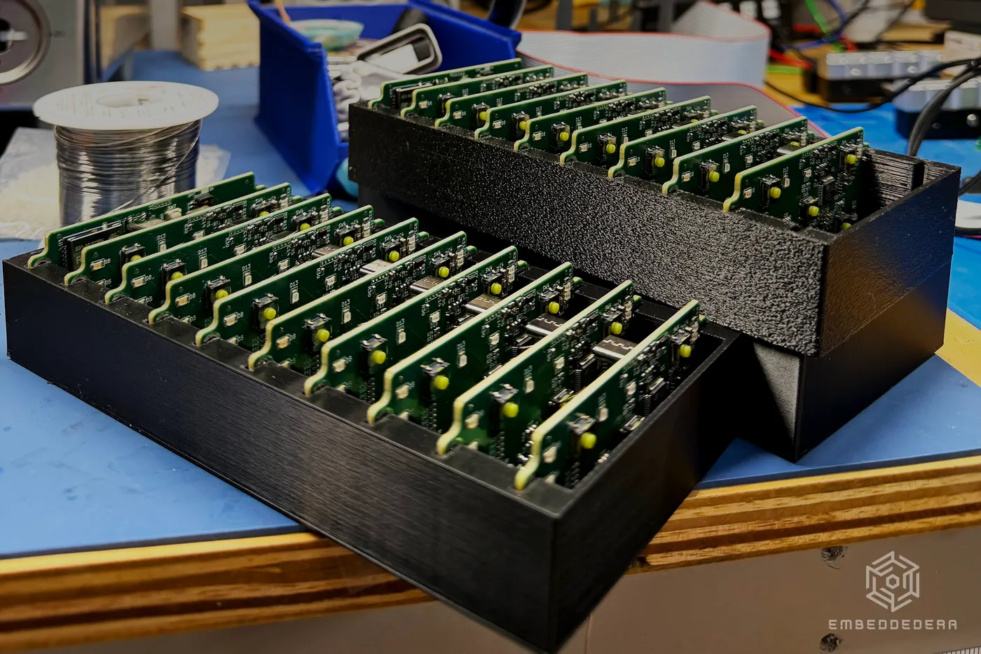 Two 3D-printed trays holding about twenty finished EmbeddedEra boards on the shop bench, test instruments behind them.
