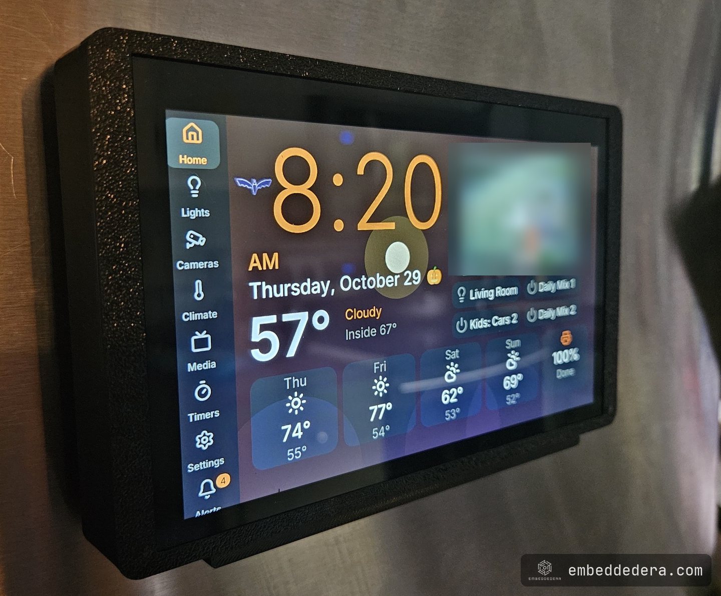 The 7-inch panel in its black printed case on a stainless fridge door, showing the home screen: clock, outside and inside temperature, a five-day forecast, quick buttons and a printer status tile.
