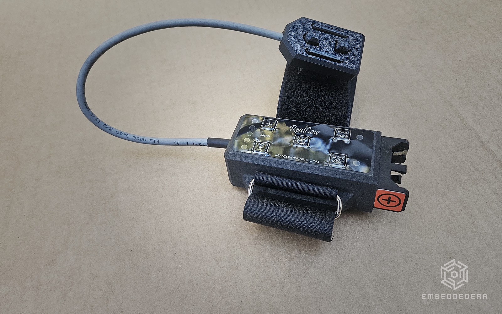 The RealCow rider's remote on a workbench: a black finger control with steering buttons, joined by a grey cable to a wrist unit with speed, power, straight mode and slow mode buttons and a hook-and-loop strap.